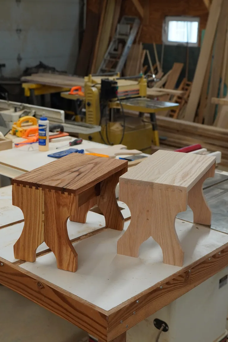 Dark oak and natural oak dovetail stools side by side on the Taylor & Grain workbench, with the shop softly behind them